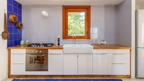 Una cucina con piano cottura a gas, forno, lavello e mobili bianchi. Si vede del verde attraverso una finestra.