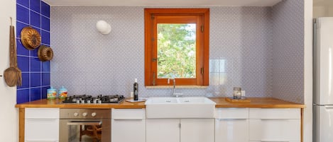 Cocina con estufa a gas, horno, pileta y muebles blancos. Se ve un ventanal con vista a vegetación.