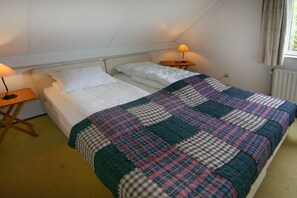 A single bed with a white duvet and a plaid blanket, two bedside tables with lamps, and a window with curtains. - Holiday home with 1 bedroom (Cadzand-Bad)
