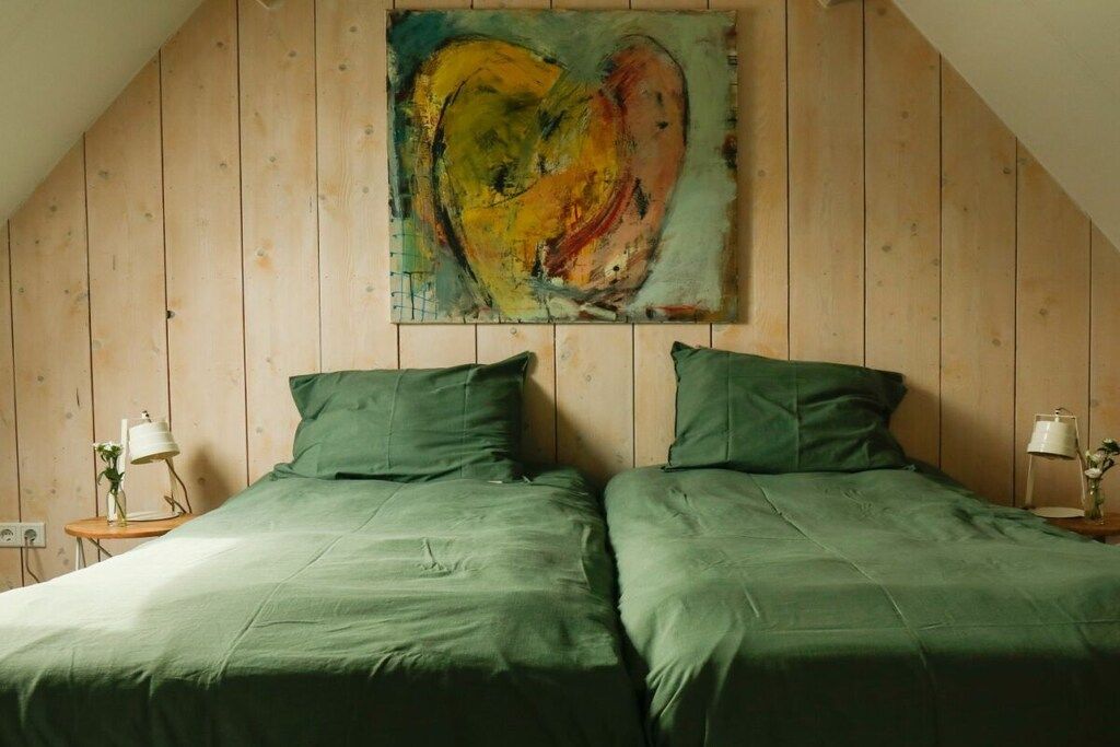 A room with a sloped ceiling and wooden walls, featuring two beds with green bedding and a large abstract painting on the wall.