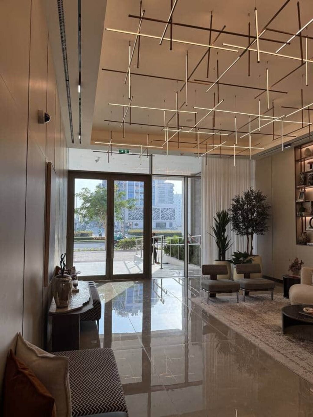 A modern hotel lobby with a glass door leading outside, a seating area with sofas and a coffee table, and a ceiling with geometric light fixtures.