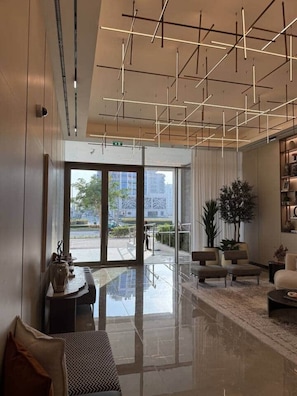 A modern hotel lobby with a glass door leading outside, a seating area with sofas and a coffee table, and a ceiling with geometric light fixtures. - Central Studio at Hyde Walk   Rooftop Pool & Free Parking (Dubai)