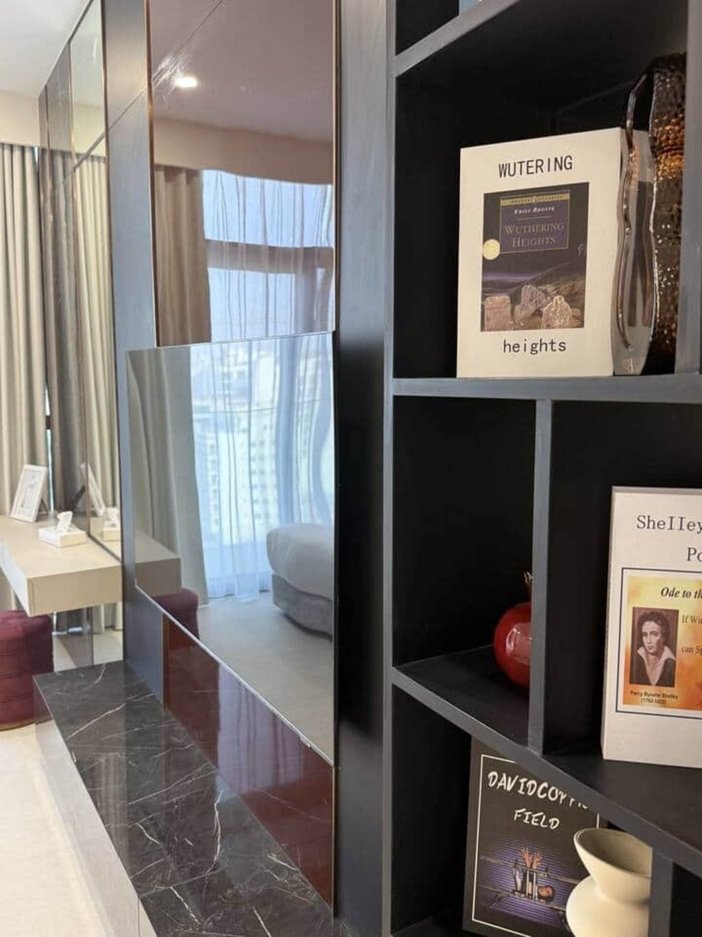 Een hotelkamer met een grote spiegel, een bed, een bureau met een stoel, en een plank met boeken en decoratieve objecten.