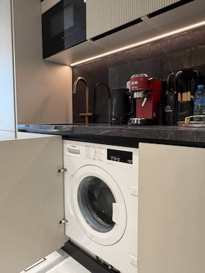 A compact kitchen with a built-in washer and dryer, a coffee maker, and a microwave. - Central Studio at Hyde Walk   Rooftop Pool & Free Parking (Dubai)
