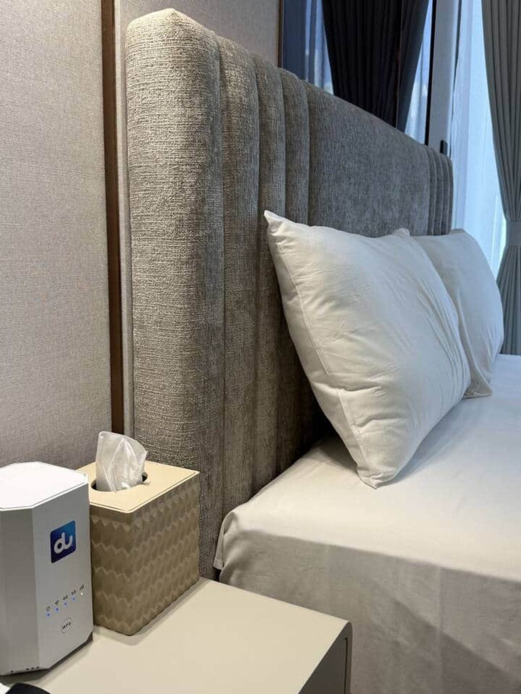 Quarto de hotel com cama arrumada, mesa de cabeceira com caixa de lenços e abajur.