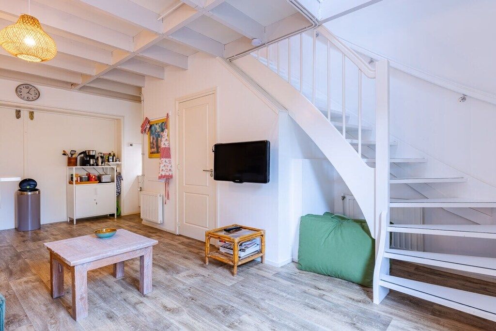 A room with a wooden table, a small shelf with books, a television on the wall, and a staircase.