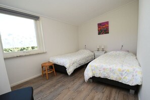 Una camera da letto con due letti singoli, uno sgabello, un comodino con una lampada e una finestra vista sul verde.