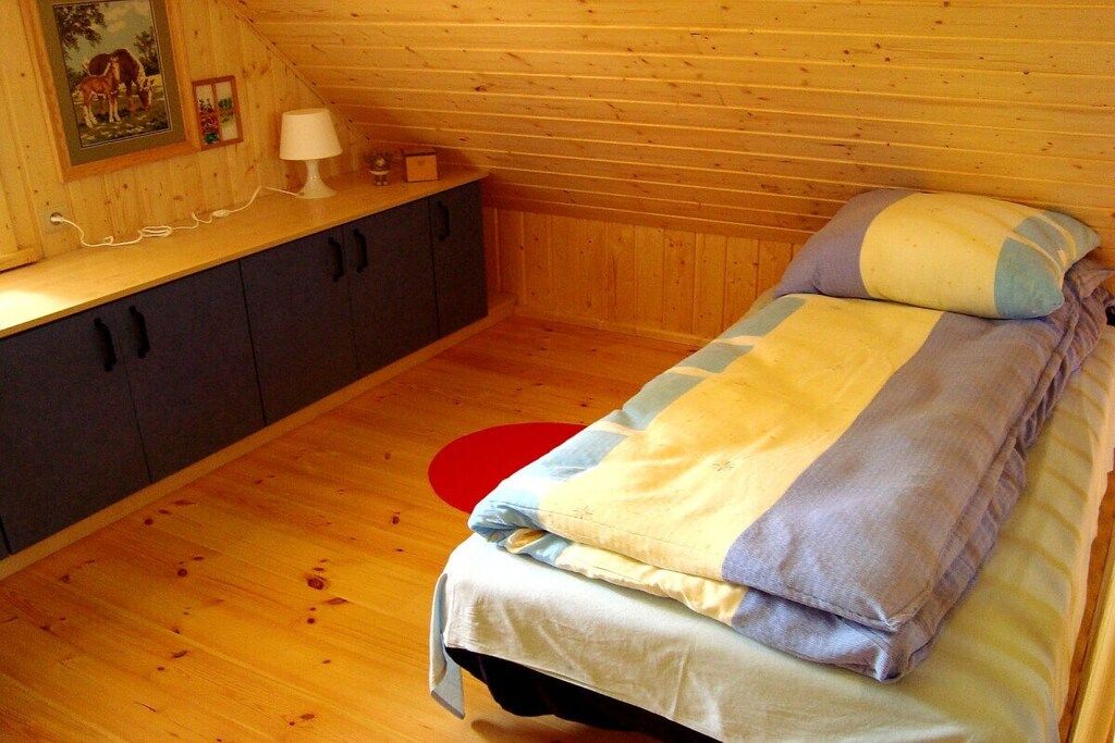 Una cama individual con una colcha y almohadas a rayas, una mesita de noche con una lámpara, un cuadro enmarcado y una alfombra roja circular sobre un suelo de madera.