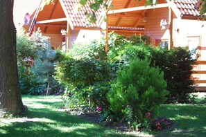 A wooden cabin with a covered porch, surrounded by lush greenery and flowering plants. - Holiday home with 1 bedroom (Dźwirzyno)