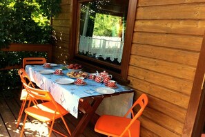 Ein Holztisch im Freien mit einer blauen Tischdecke, umgeben von orangefarbenen Stühlen, vor einer Holzwand mit einem Fenster.