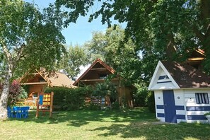 Eine Holzhütte mit geneigtem Dach, umgeben von üppigem Grün und einem kleinen Spielbereich mit blauen Stühlen.