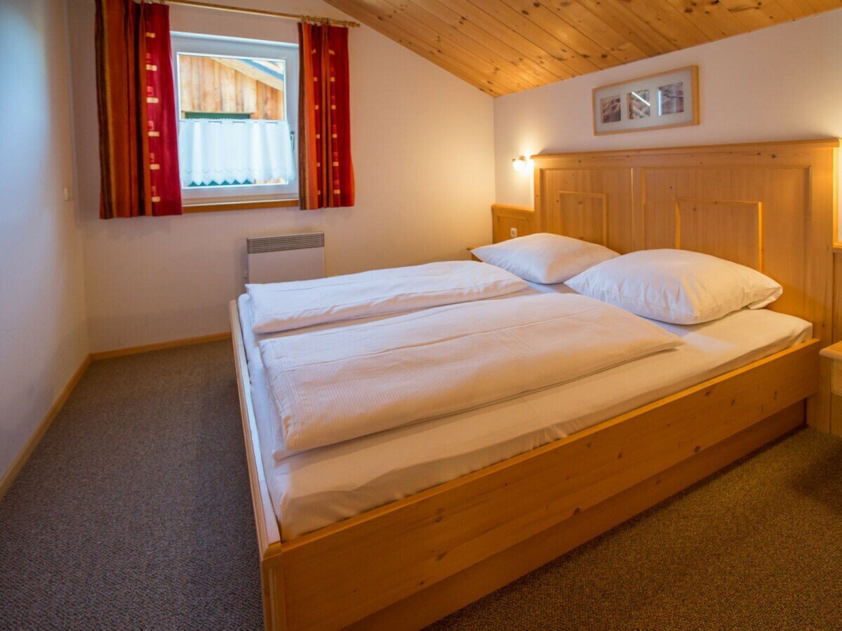 Un lit double avec du linge de lit blanc, une tête de lit en bois, une table de chevet avec une lampe et une fenêtre avec des rideaux.