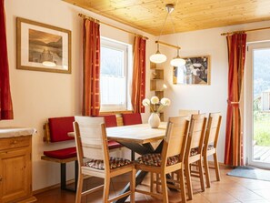 Een eethoek met een houten tafel en stoelen, rode kussens, een vaas met bloemen en uitzicht op groen buiten.