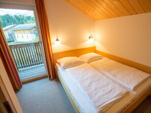 Quarto com teto de madeira, uma cama de casal, duas luminárias fixadas na parede e uma varanda com vista para casas e árvores.