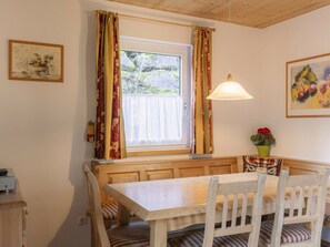 Uma área de refeições com mesa de madeira e bancos, janela com cortinas e luminária pendente.