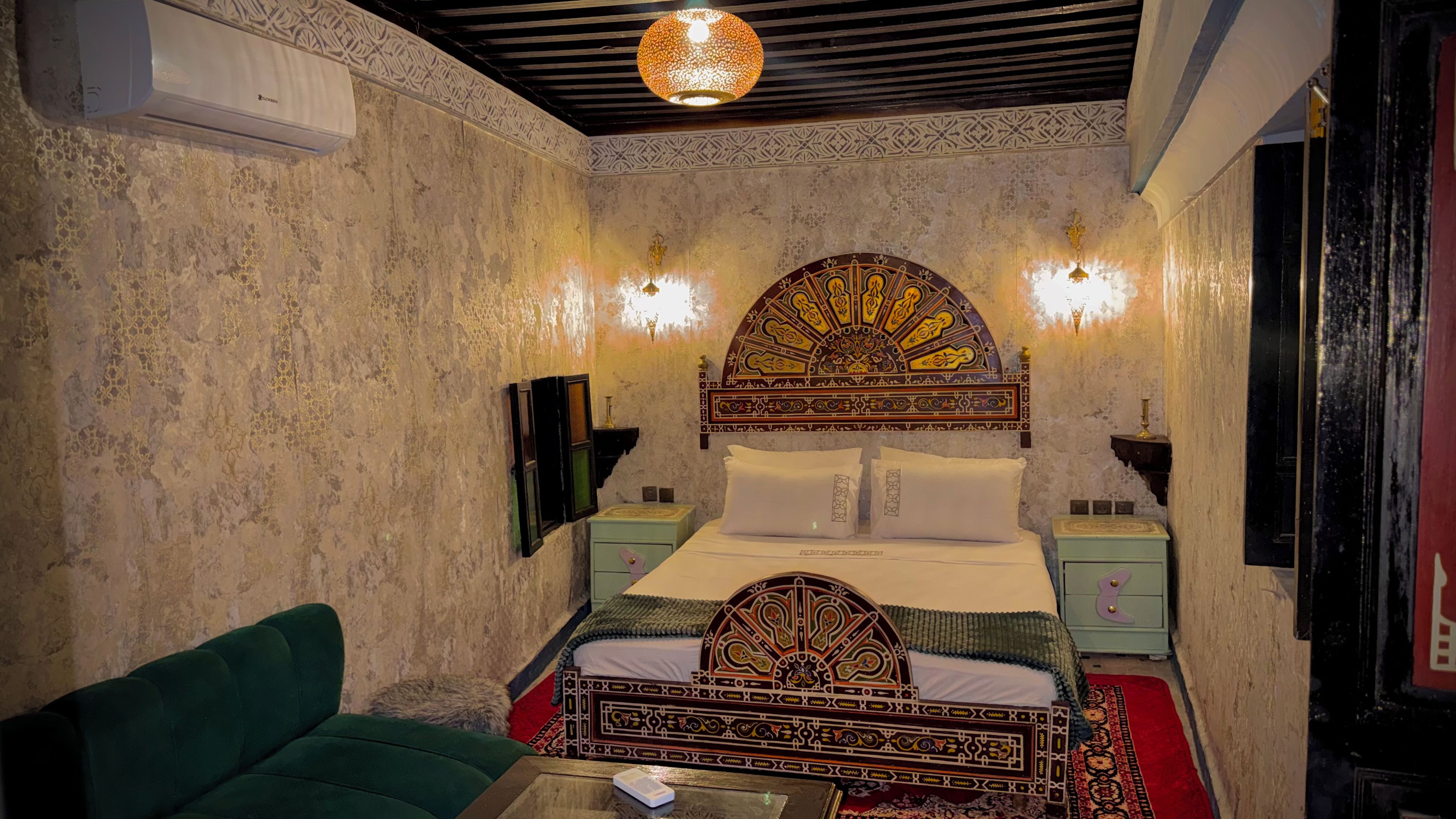 Una habitación con cama, sofá verde, cabecero decorativo, lámparas de pared y una lámpara en el techo.