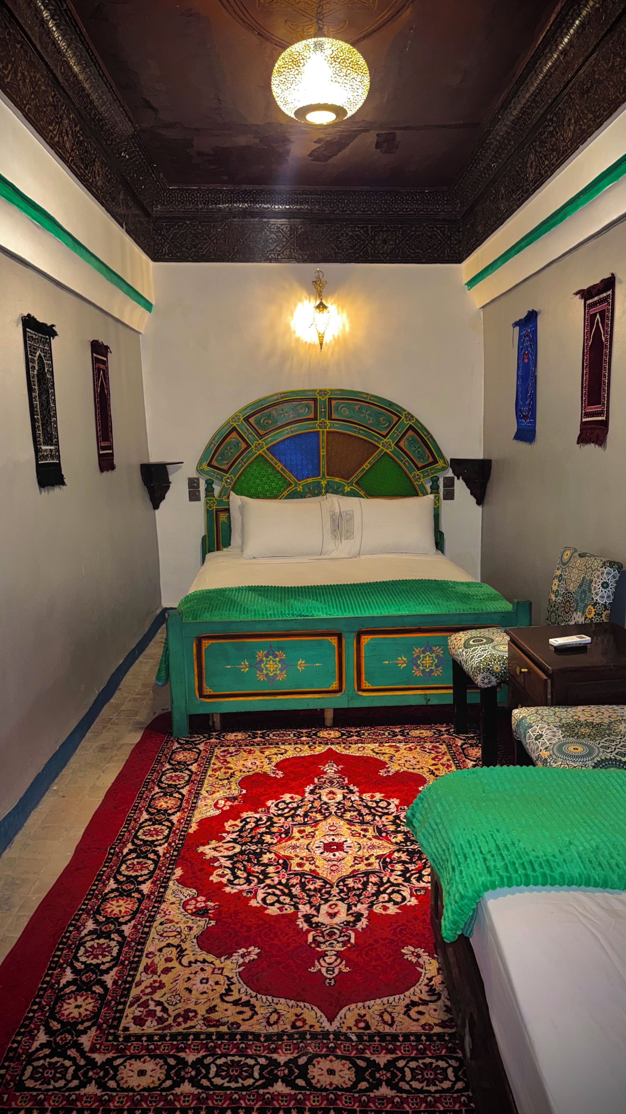 Una habitación con una cama, una mesita de noche, una lámpara, un tapete estampado y un cabecero decorativo.