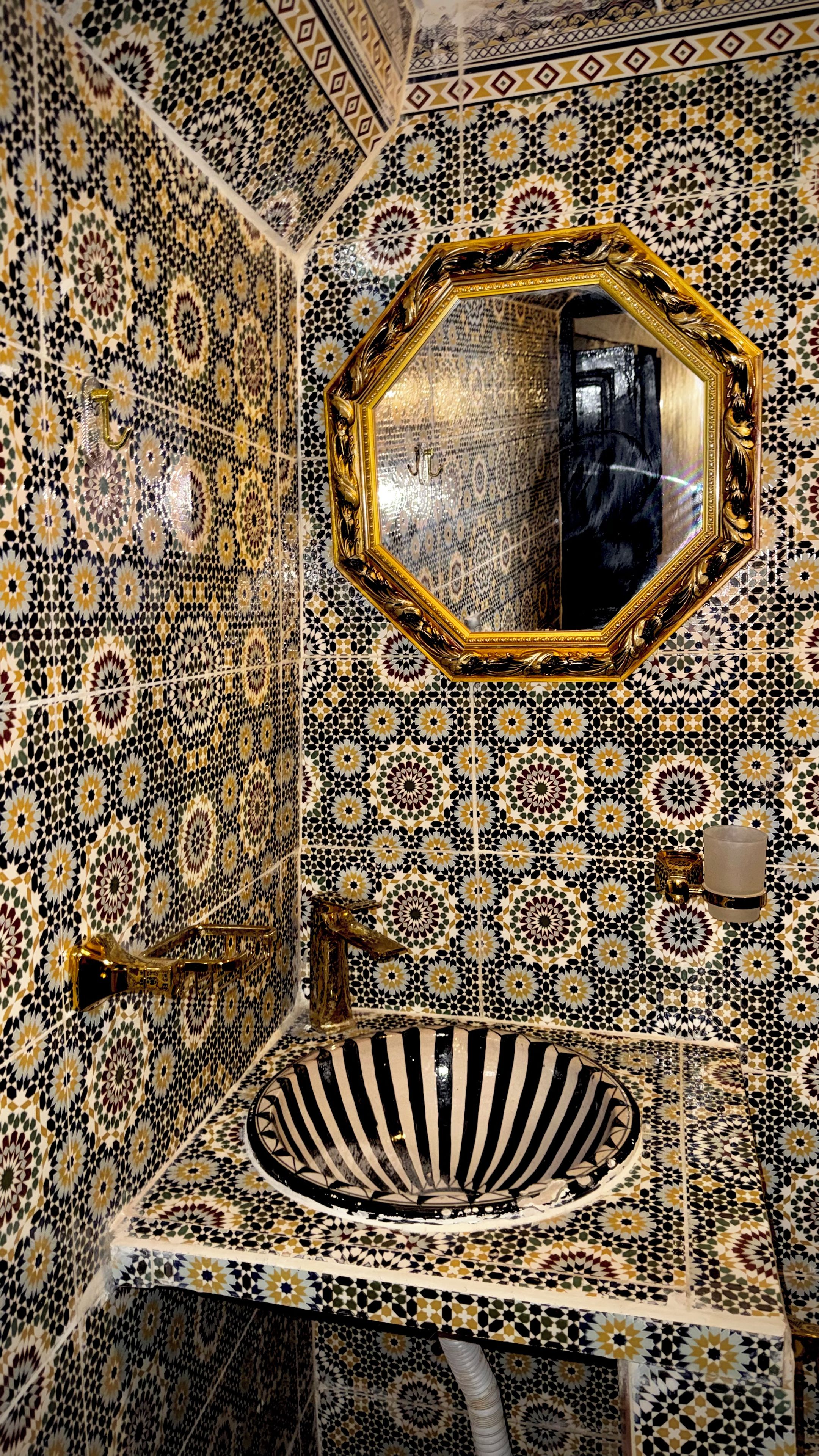 Un baño con pared decorada con azulejos, un lavamanos con lavabo a rayas negras y blancas, y un espejo con marco dorado.