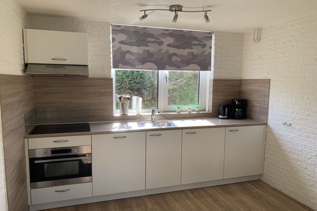 Een moderne keuken met witte kastjes, een inbouwoven, een roestvrijstalen spoelbak en een raam met een camouflage jaloezie.