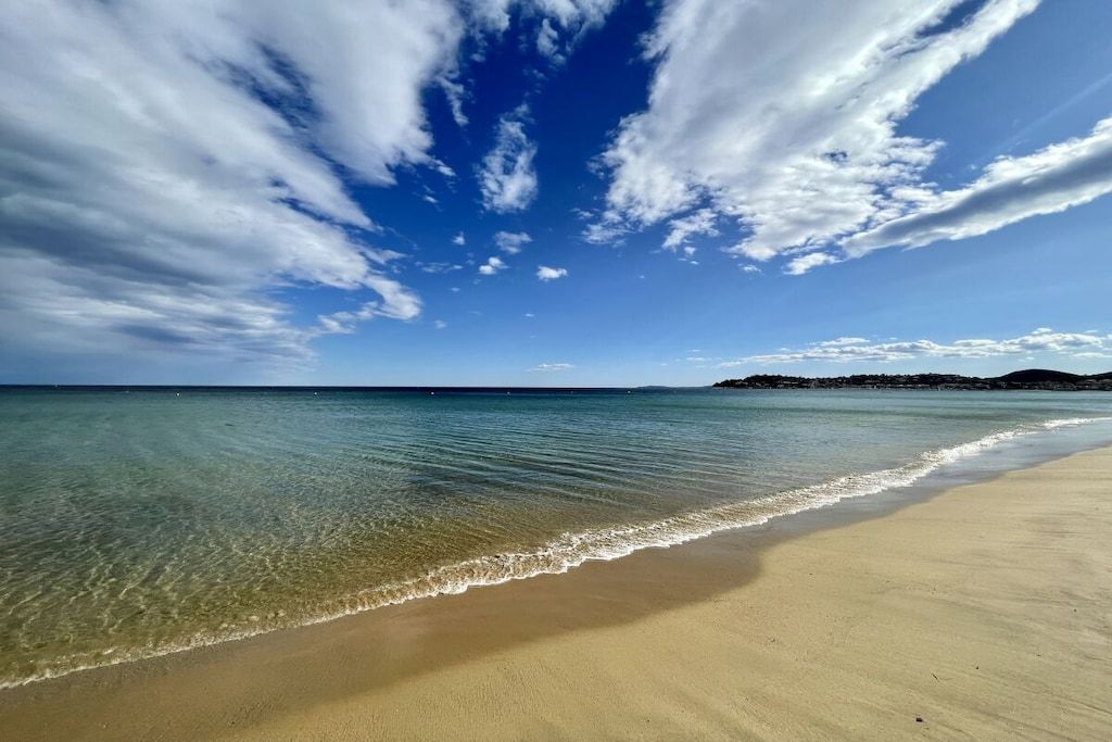 Une plage à l’eau claire, un rivage sablonneux et une terre lointaine sous un ciel bleu parsemé de nuages.