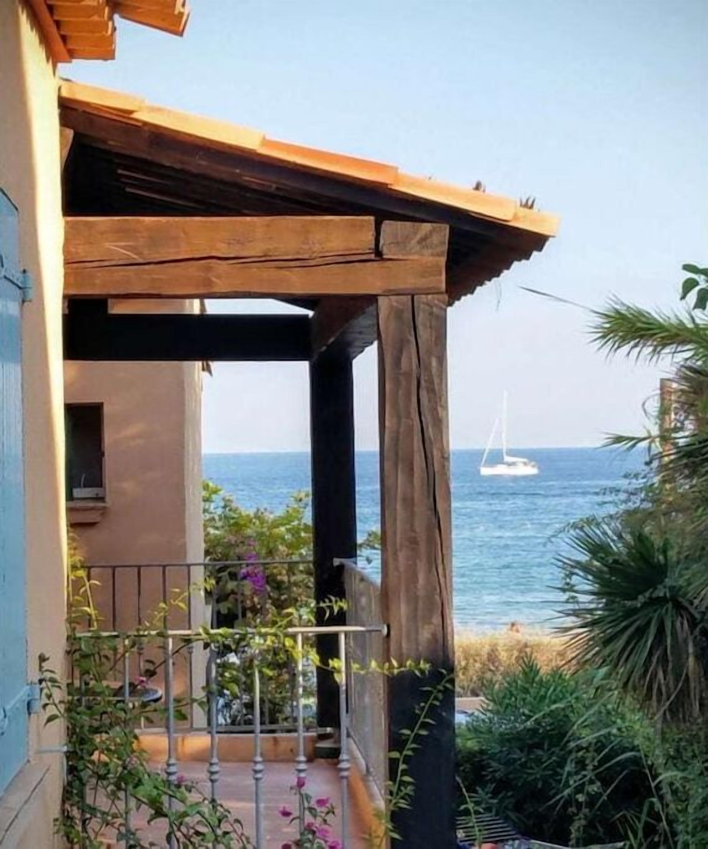 Een balkon met een houten pergola, een hekwerk en uitzicht op zee met een zeilboot.