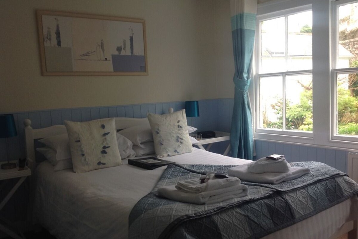 A neatly made bed with a blue and white bedspread, a bedside table with a lamp, and a window with curtains.