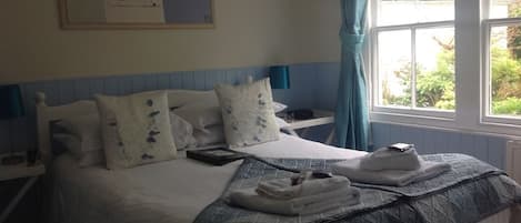 Una cama bien hecha con colcha azul y blanca, una mesita de noche con lámpara y una ventana con cortinas.