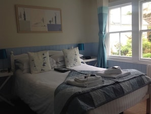 A neatly made bed with a blue and white bedspread, a bedside table with a lamp, and a window with curtains.