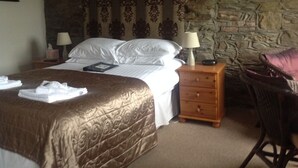 A hotel room with a large bed, a wooden nightstand, a chair, and a stone wall.