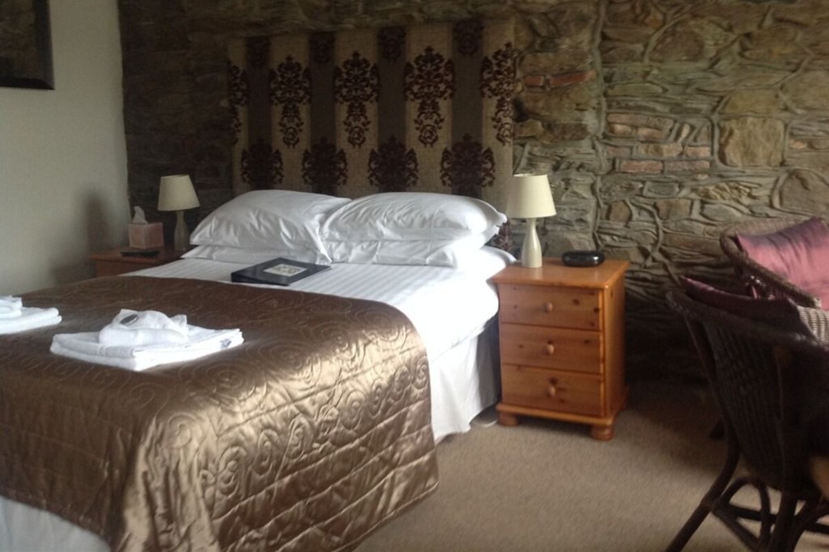 A hotel room with a large bed, a wooden nightstand, a chair, and a stone wall.