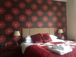Habitación de hotel con papel tapiz floral, una cama con ropa de cama roja, dos lámparas de noche y una cama bien hecha con toallas.