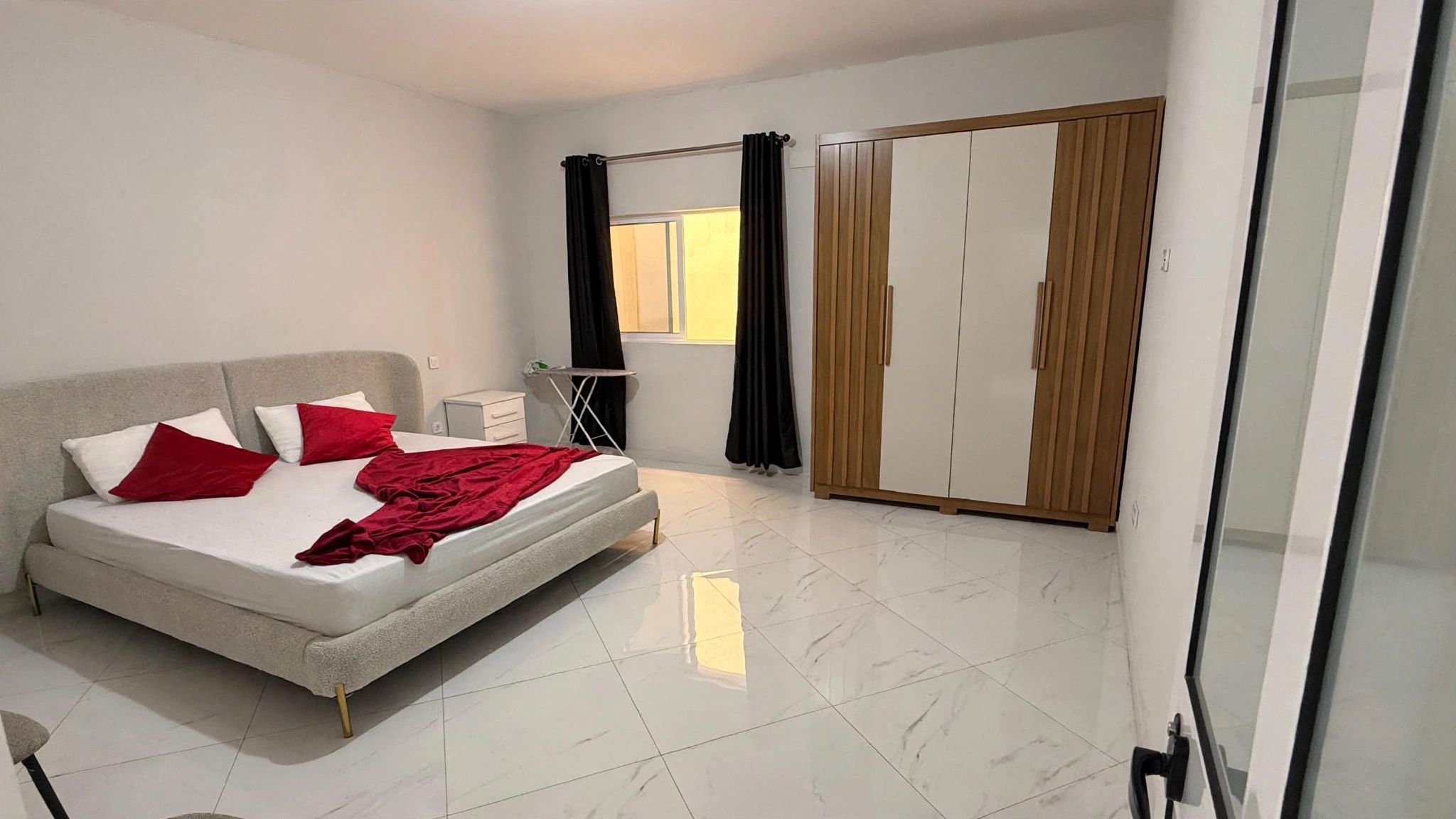 Ein Schlafzimmer mit einem Bett, einem Kleiderschrank, einem Fenster mit Vorhängen und einem Fliesenboden.