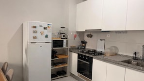Kompakti keittiö, jossa on valkoiset kaapit, valkoinen jääkaappi, mikroaaltouuni, kaasuliesi ja erilaisia keittiövälineitä.