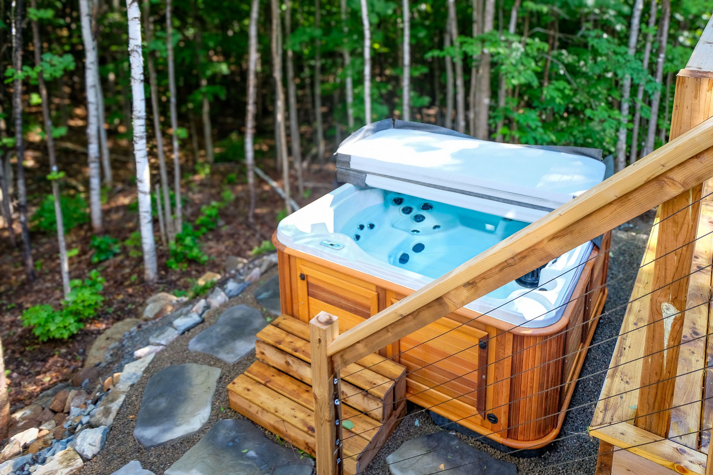 A hot tub with a wooden exterior, surrounded by a stone pathway and a wooden fence, set in a forested area.