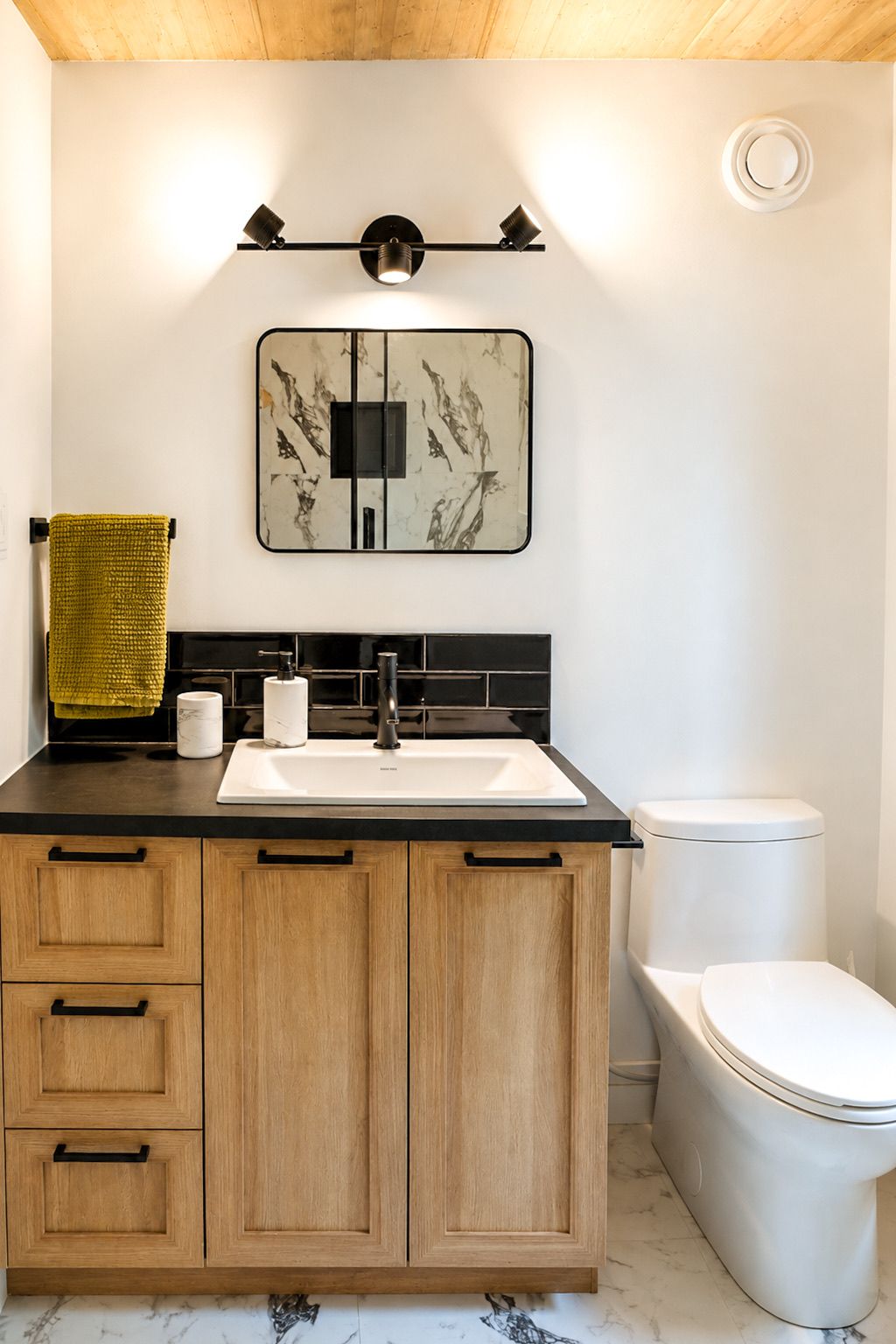 A modern bathroom with a black countertop, wooden cabinets, a white toilet, and a wall-mounted light fixture.