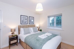 A bedroom with a bed, bedside lamps, a window with blinds, and two framed pictures on the wall. - 2-bed Apartment in Manchester - Free Parking (Altrincham)