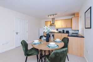 A modern kitchen with wooden cabinets, a gas stove, and a dining area with a round table and green upholstered chairs. - 2-bed Apartment in Manchester - Free Parking (Altrincham)