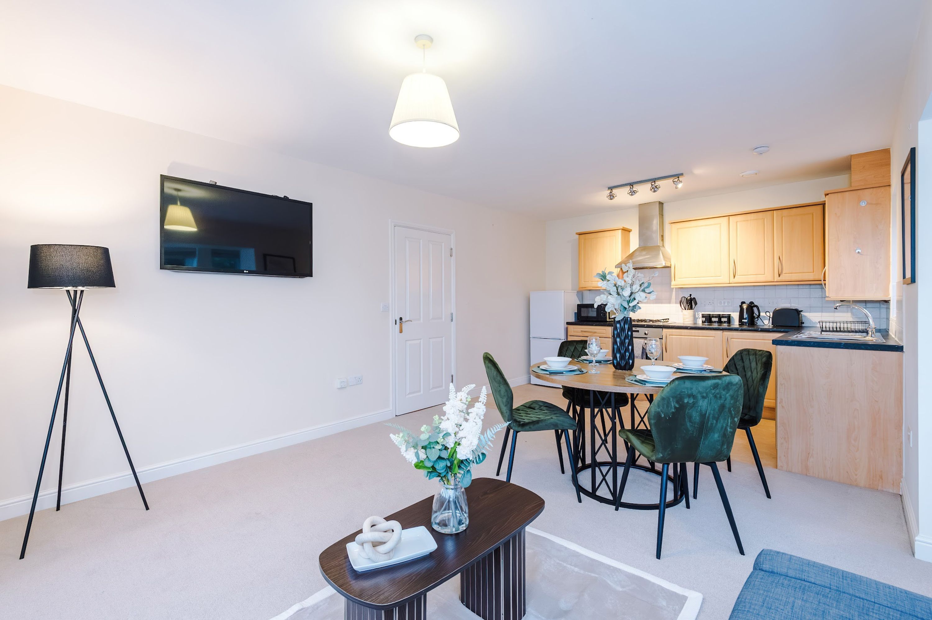 A modern kitchen with wooden cabinets, a dining area with a round table and chairs, a flat-screen TV mounted on the wall, and a floor lamp.