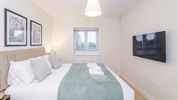 A bedroom with a bed, a TV mounted on the wall, a window with blinds, and two framed pictures on the wall.