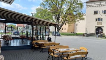 Un espacio al aire libre con mesas y bancos de madera, una zona cubierta con sillas y un árbol grande al fondo.