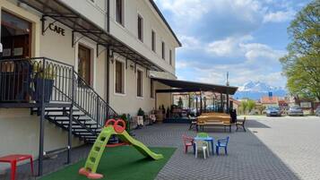 Un edificio con letrero de cafetería, mesas al aire libre, una zona de juegos para niños y un patio empedrado.