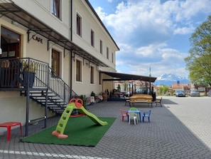 A building with a cafe sign, outdoor seating, a children's play area, and a paved courtyard. - Penzión Berg (Kezmarok)