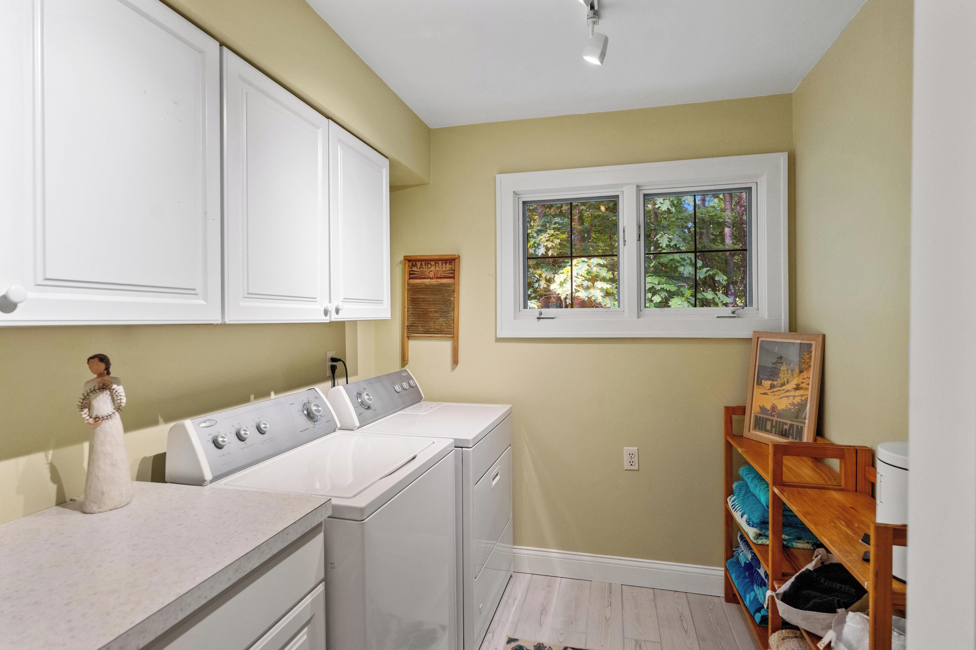 A laundry room with a washer and dryer, a wooden shelf with folded clothes, and a window with a view of trees.