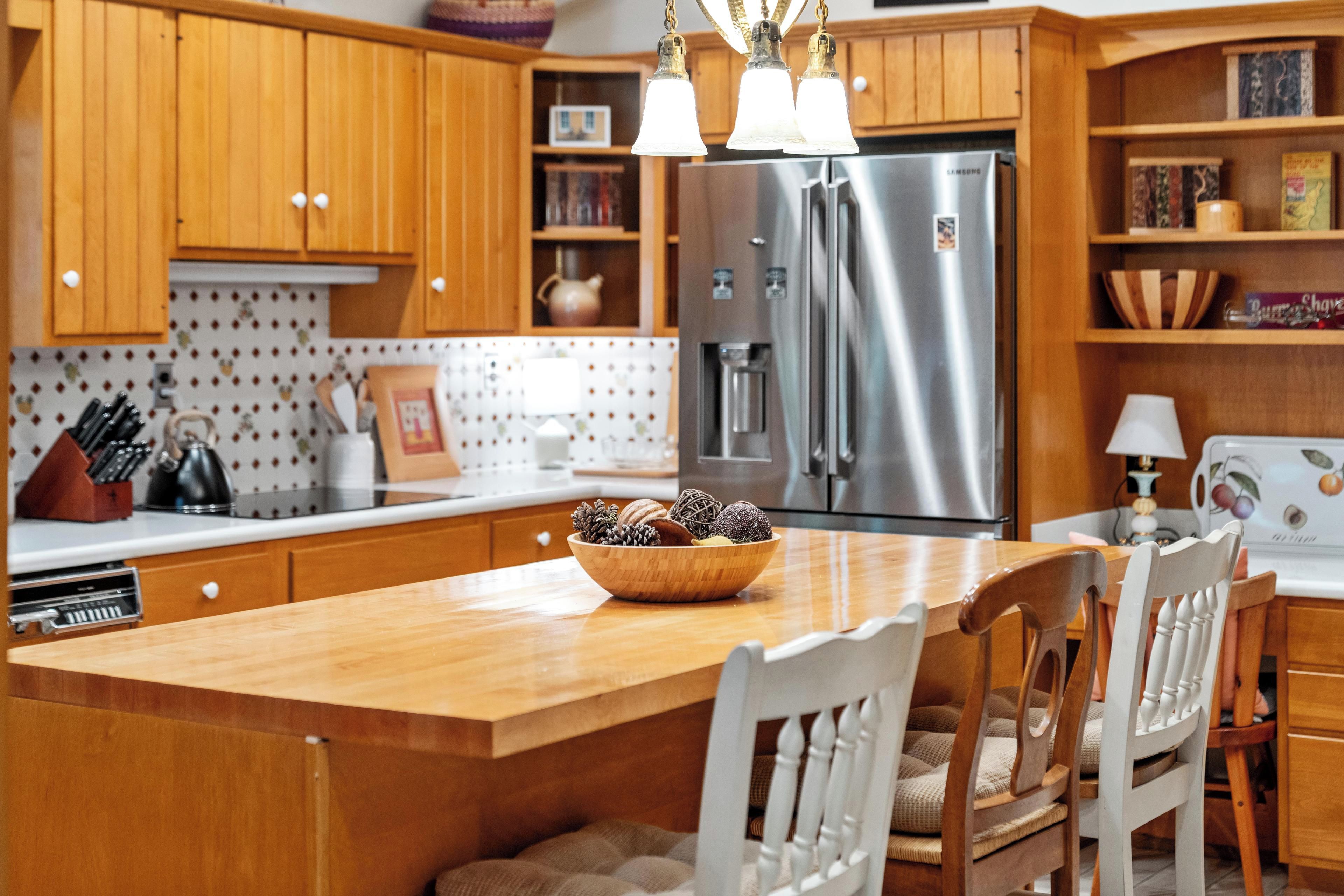 Una cocina con gabinetes de madera, un refrigerador de acero inoxidable, una mesa de comedor de madera con sillas, y un tazón de bocadillos.