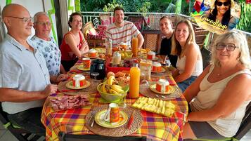Un grupo de personas sentadas en una mesa al aire libre, con diversa comida y bebidas, rodeadas de follaje.