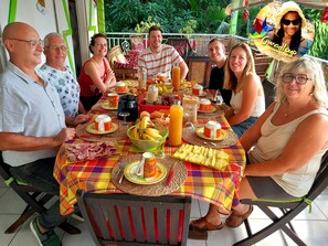 A group of people seated at an outdoor table with a variety of food and drinks, surrounded by greenery.