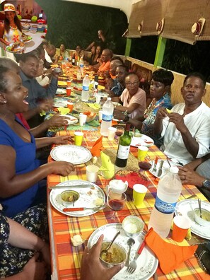 Un groupe de personnes est assis autour d’une table, devant des assiettes, des tasses et des bouteilles, ce qui laisse supposer qu’il s’agit d’un repas partagé.