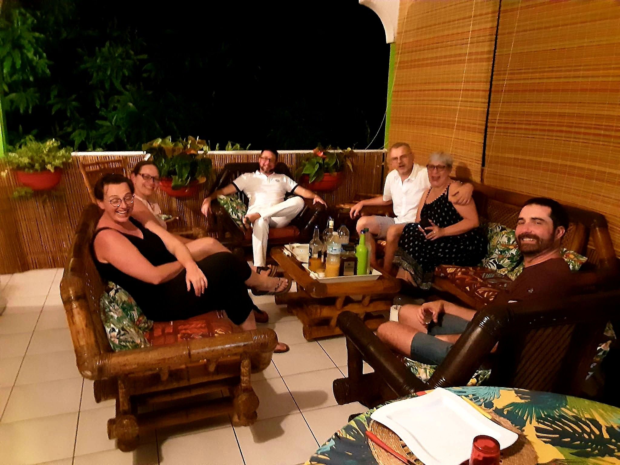 A group of people seated on outdoor furniture around a table with drinks, in a setting with plants and a wooden backdrop.