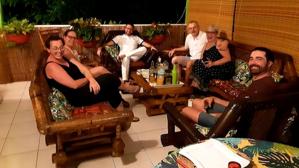 A group of people seated on outdoor furniture around a table with drinks, in a setting with plants and a wooden backdrop.