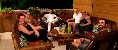 Un groupe de personnes est assis sur des meubles d’extérieur autour d’une table où se trouvent des boissons, dans un décor agrémenté de plantes et d’un fond en bois.